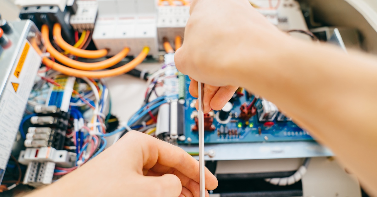Hands use a screwdriver to adjust internal components inside an open electronic device with visible wiring.