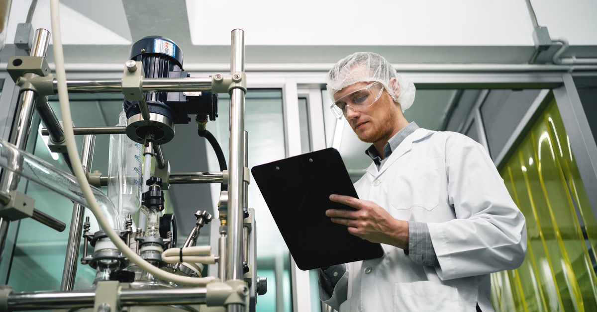 A technician in protective gear reviews notes on a clipboard beside industrial laboratory equipment.
