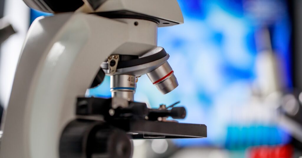 Close-up of laboratory microscope with objective lenses focused on slide, blurred scientific equipment in background.