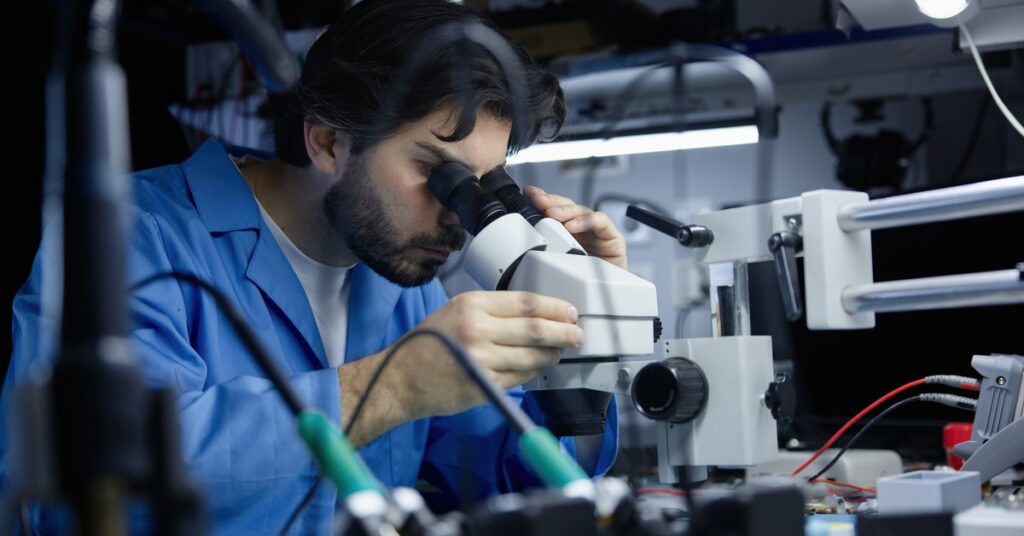 A technician is using a microscope to examine components in electronics lab with tools, wires, and testing equipment.