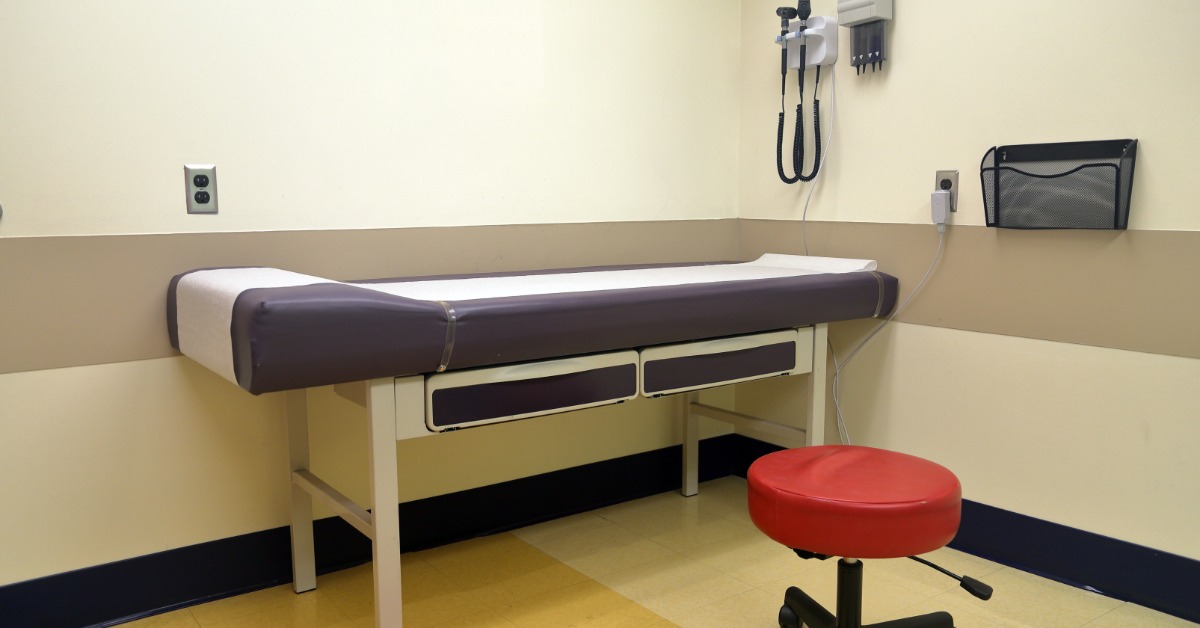An empty medical examination room with a padded exam table, rolling stool, and diagnostic tools mounted on the wall.