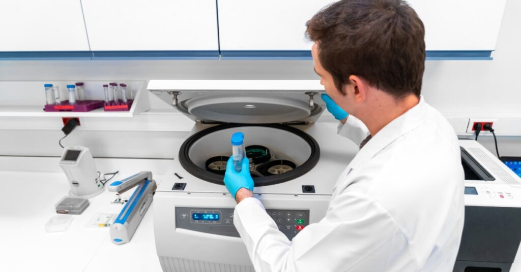 A laboratory technician places a sample tube into a centrifuge machine while preparing specimens for testing.
